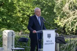 Mężczyzna w garniturze przemawia przy mównicy z napisem WILLA DECJUSZA.