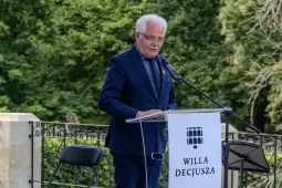 Mężczyzna w garniturze przemawia przy mównicy z napisem WILLA DECJUSZA. W tle zieleń drzew.