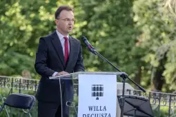 Mężczyzna w garniturze przemawia przy mikrofonie, w tle zieleń drzew.