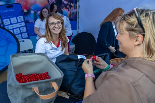 Kobieta w okularach uśmiecha się, rozmawiając z inną osobą przy stoisku.