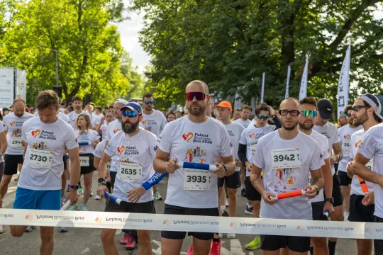 Biegacze w koszulkach startowych przygotowują się do biegu, trzymając w rękach pałeczki.