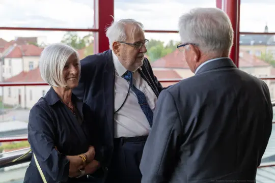 Trzy osoby rozmawiają w eleganckich strojach, w tle widoczny krajobraz miejski.