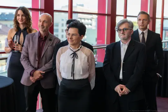 Grupa elegancko ubranych ludzi na przyjęciu, stojących w sali z dużymi oknami.