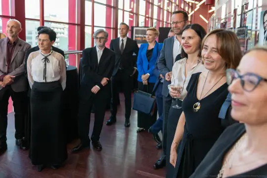 Grupa ludzi w eleganckich strojach, uśmiechających się i rozmawiających w nowoczesnym wnętrzu.