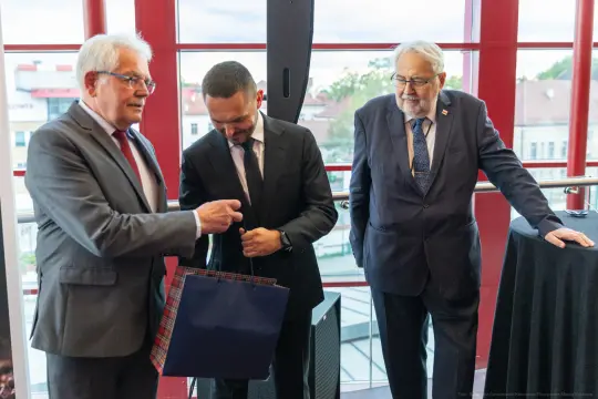 Trzech mężczyzn w formalnych strojach, rozmawiających przy stole w eleganckim wnętrzu.
