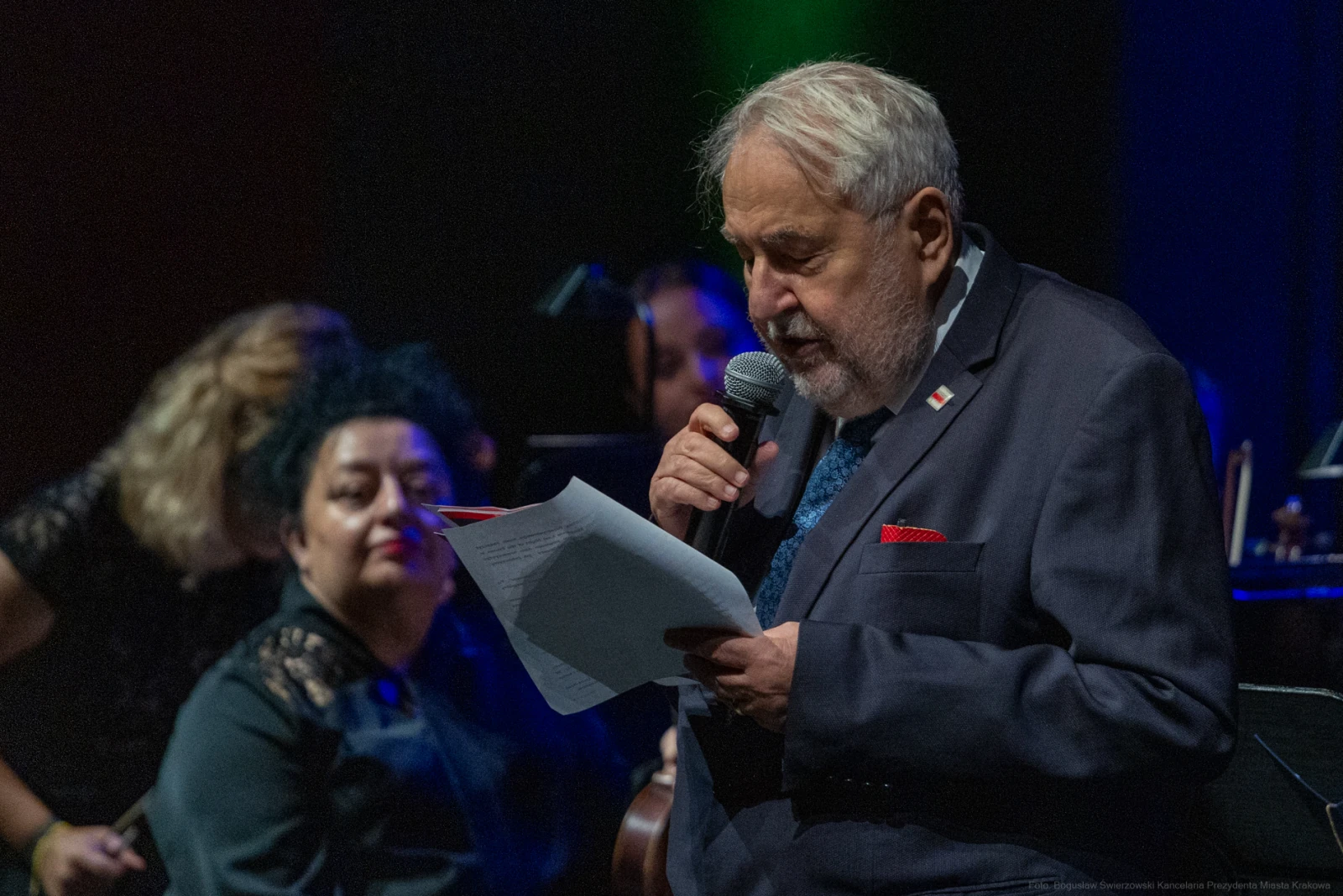 Mężczyzna w garniturze czyta tekst, w tle muzycy i kobieta z ciemnymi włosami.  Autor: Bogusław Świerzowski