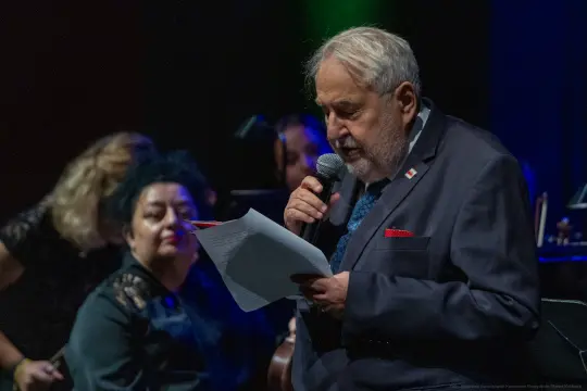 Mężczyzna w garniturze czyta tekst, w tle muzycy i kobieta z ciemnymi włosami.
