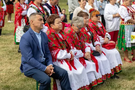 Grupa osób w tradycyjnych strojach ludowych siedzących na trawie podczas wydarzenia.