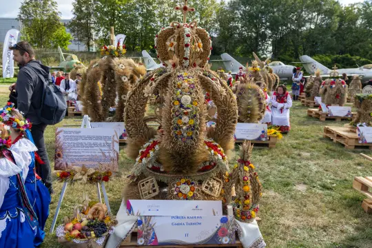 Wieniec dożynkowy ozdobiony kwiatami i zbożem, otoczony ludźmi w tradycyjnych strojach.