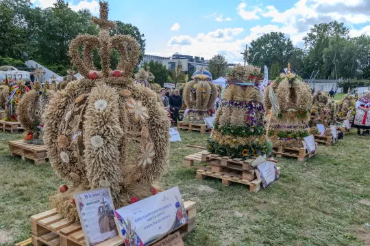 Wieniec dożynkowy w kształcie korony, ozdobiony kwiatami i zbożem, na tle festiwalu.