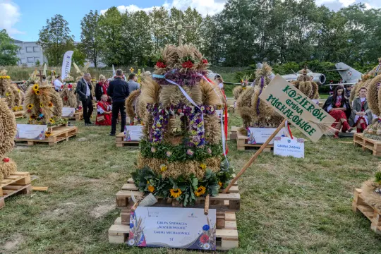 Wieniec dożynkowy ozdobiony kwiatami i zbożem, z tabliczką informacyjną.