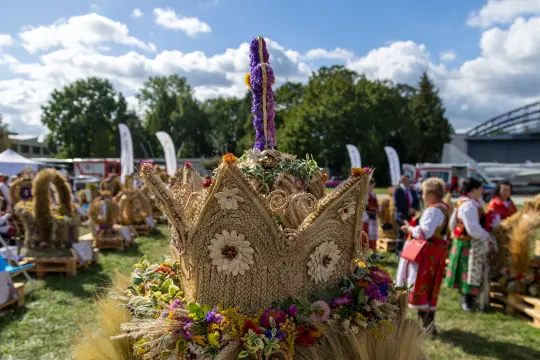 Wieniec dożynkowy ozdobiony kwiatami i zbożem, w tle festiwal z ludźmi w tradycyjnych strojach.