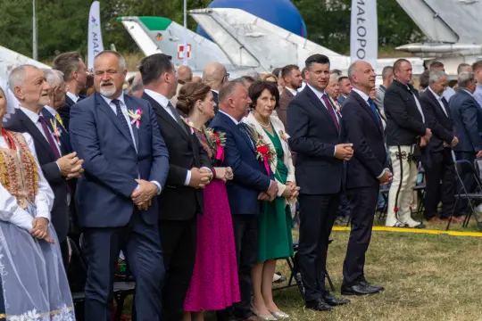 Grupa ludzi w eleganckich strojach, niektórzy w tradycyjnych, podczas uroczystości na świeżym powietrzu.
