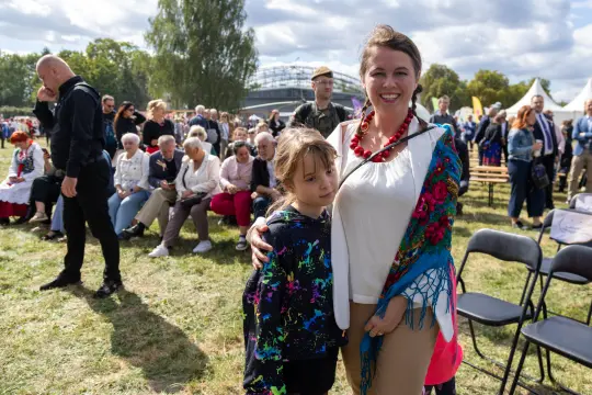 Kobieta w kolorowej chustce z dzieckiem, w tle tłum ludzi na wydarzeniu plenerowym.