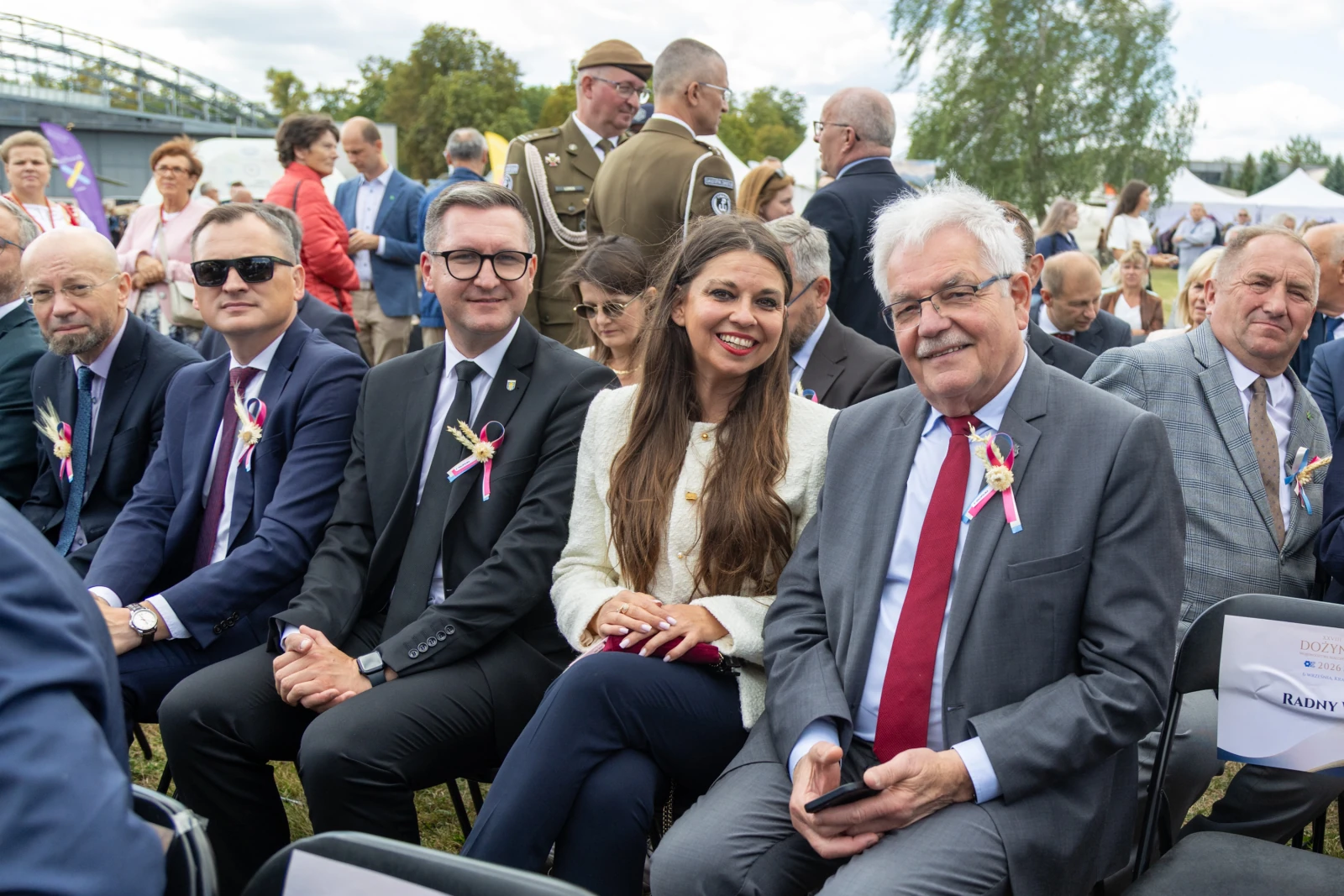 Grupa osób w garniturach siedząca na wydarzeniu plenerowym, z uśmiechami i odznaczeniami na ubraniach.  Autor: Bogusław Świerzowski