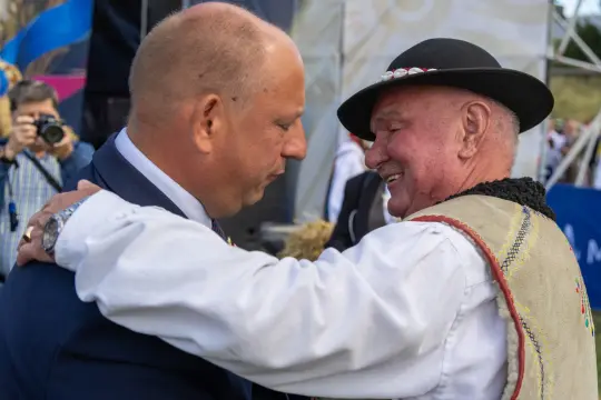 Uścisk dłoni dwóch mężczyzn w tradycyjnych strojach, w tle tłum i scena.