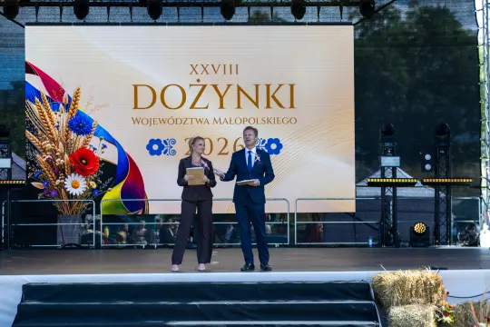 Ceremonia dożynkowa z prowadzącymi na scenie, dekoracje i logo województwa Małopolskiego.