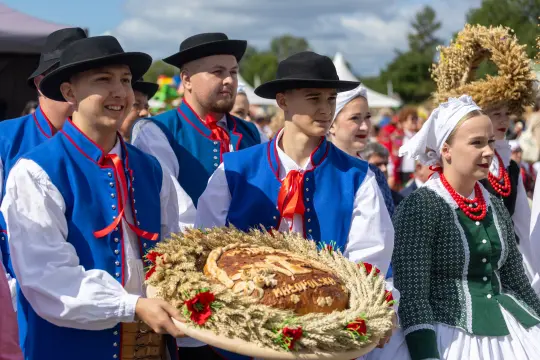 Grupa ludzi w tradycyjnych strojach ludowych niesie chleb ozdobiony zbożem.