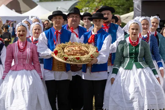 Grupa ludzi w tradycyjnych strojach ludowych z wieńcem dożynkowym.