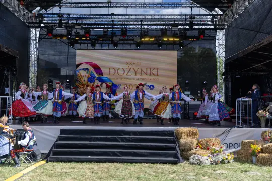 Grupa tancerzy w tradycyjnych strojach ludowych tańczy na scenie podczas dożynek.