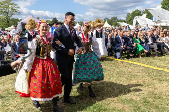 Mężczyzna w garniturze idzie w towarzystwie kobiet w tradycyjnych strojach ludowych.
