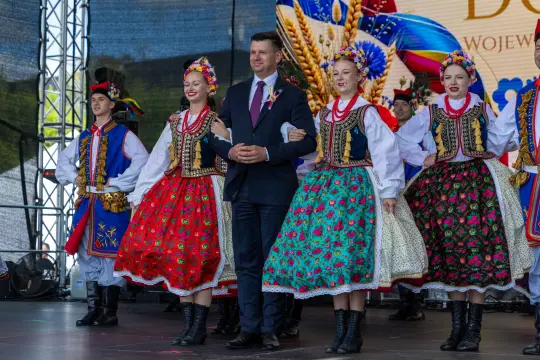 Mężczyzna w garniturze tańczy z grupą osób w tradycyjnych strojach ludowych.