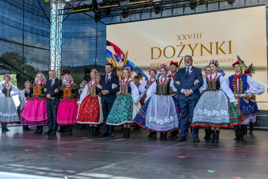 Grupa ludzi w tradycyjnych strojach ludowych tańcząca na scenie podczas dożynek.