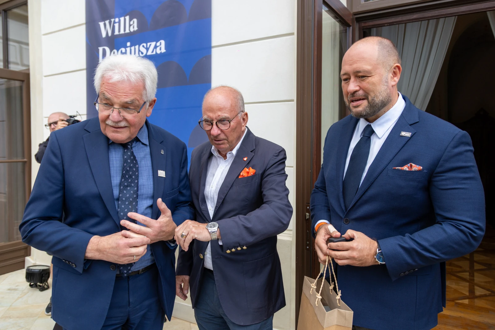 Trzech mężczyzn w eleganckich garniturach rozmawia przed budynkiem.  Autor: Bogusław Świerzowski