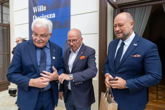 Trzech mężczyzn w eleganckich garniturach rozmawia przed budynkiem.