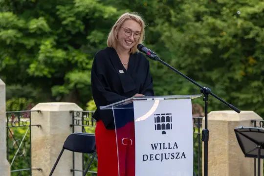 Kobieta w okularach stoi przy mównicy, uśmiechając się i przemawiając.