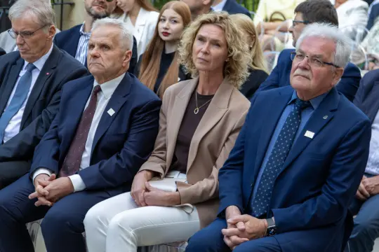Grupa osób siedzących na wydarzeniu, wśród nich kobieta w jasnym garniturze i mężczyźni w eleganckich strojach.