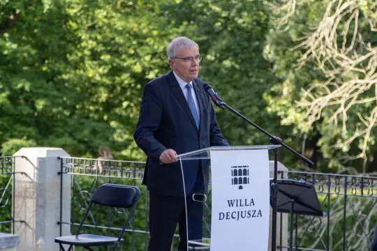 Mężczyzna w garniturze przemawia przy mównicy z napisem WILLA DECJUSZA. Tło z zielonymi drzewami.