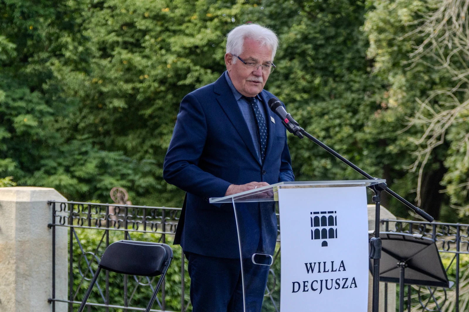 Mężczyzna w garniturze przemawia przy mównicy z napisem Willa Decjusza. W tle zieleń drzew.  Autor: Bogusław Świerzowski
