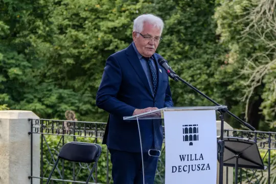 Mężczyzna w garniturze przemawia przy mównicy z napisem Willa Decjusza. W tle zieleń drzew.