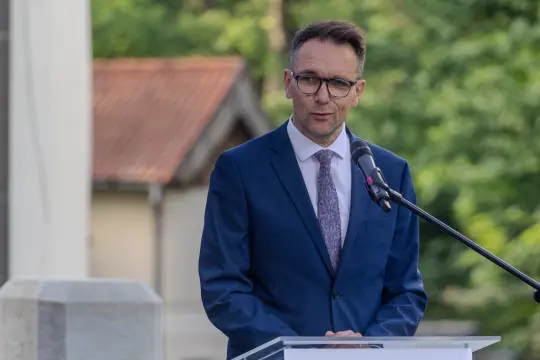 Mężczyzna w garniturze przemawia przy mównicy na tle zieleni.