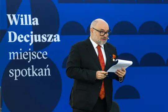 Mężczyzna w garniturze przemawia na tle niebieskiej ściany z napisem Willa Decjusza.