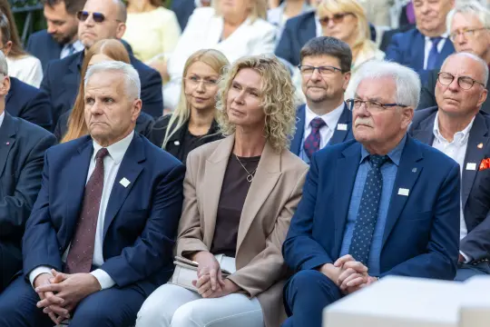 Grupa osób siedzących na wydarzeniu, w tym mężczyźni w garniturach i kobieta w jasnej bluzce.
