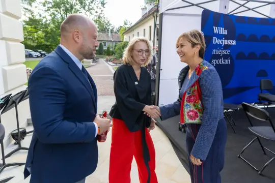 Spotkanie trzech osób w eleganckim otoczeniu, jedna z nich wita drugą.