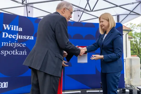 Uroczystość wręczenia dokumentu w Willi Decjusza. Mężczyzna i kobieta podają sobie ręce.