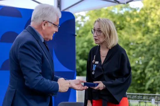 Wręczenie medalu przez mężczyznę kobiecie w eleganckich strojach, w tle zieleń.