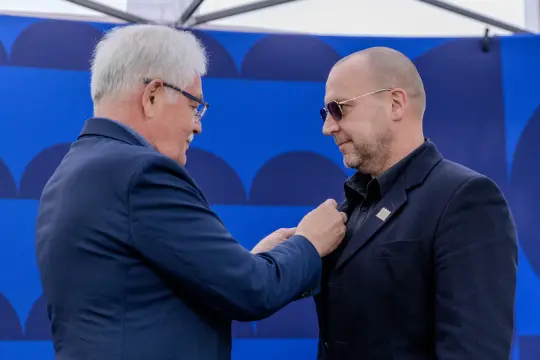 Mężczyzna w okularach przeciwsłonecznych otrzymuje odznaczenie od innego mężczyzny.