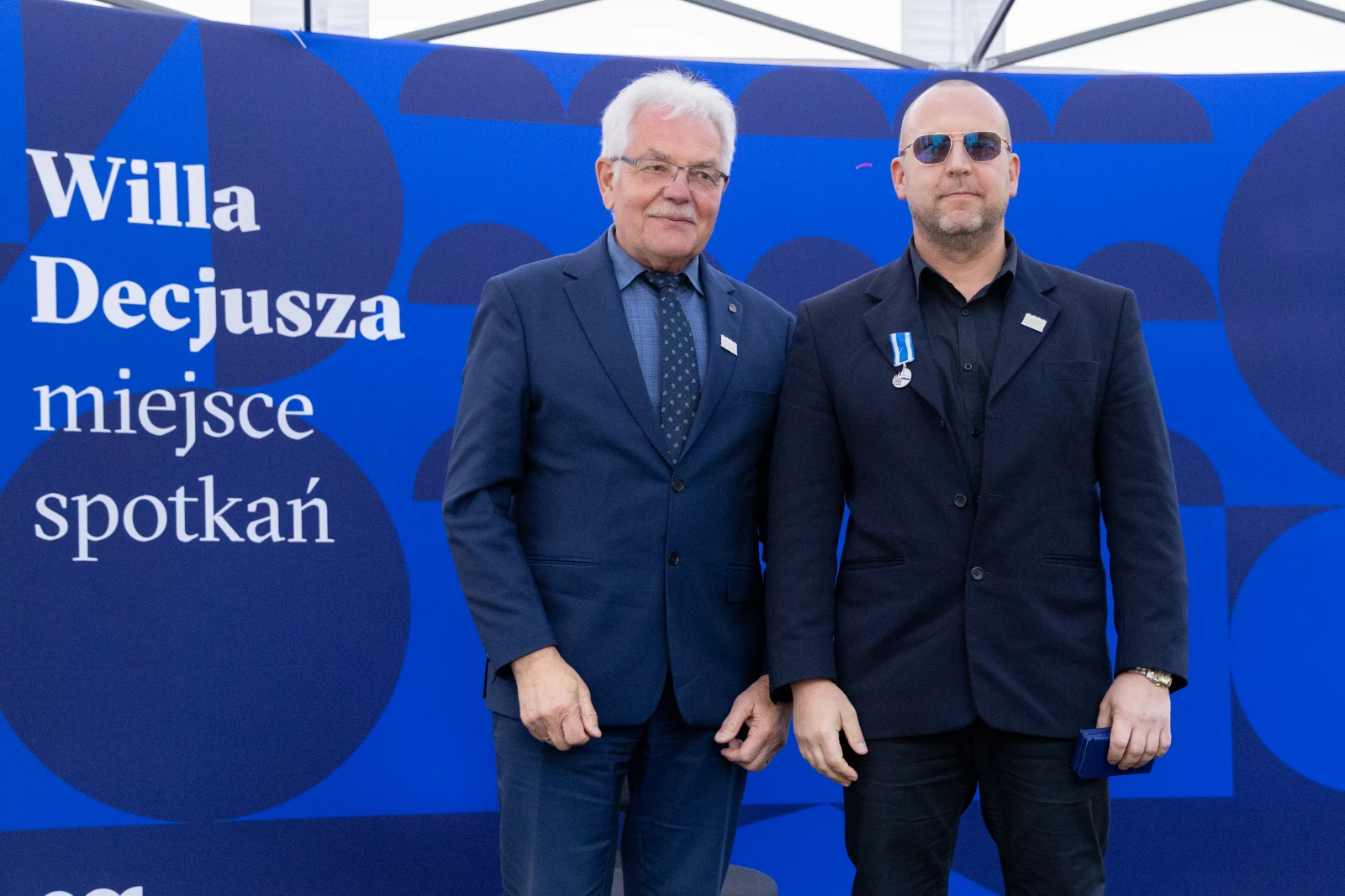 Dwaj mężczyźni stoją obok siebie, jeden z medalem, w tle niebieska ściana z napisem.  Autor: Bogusław Świerzowski
