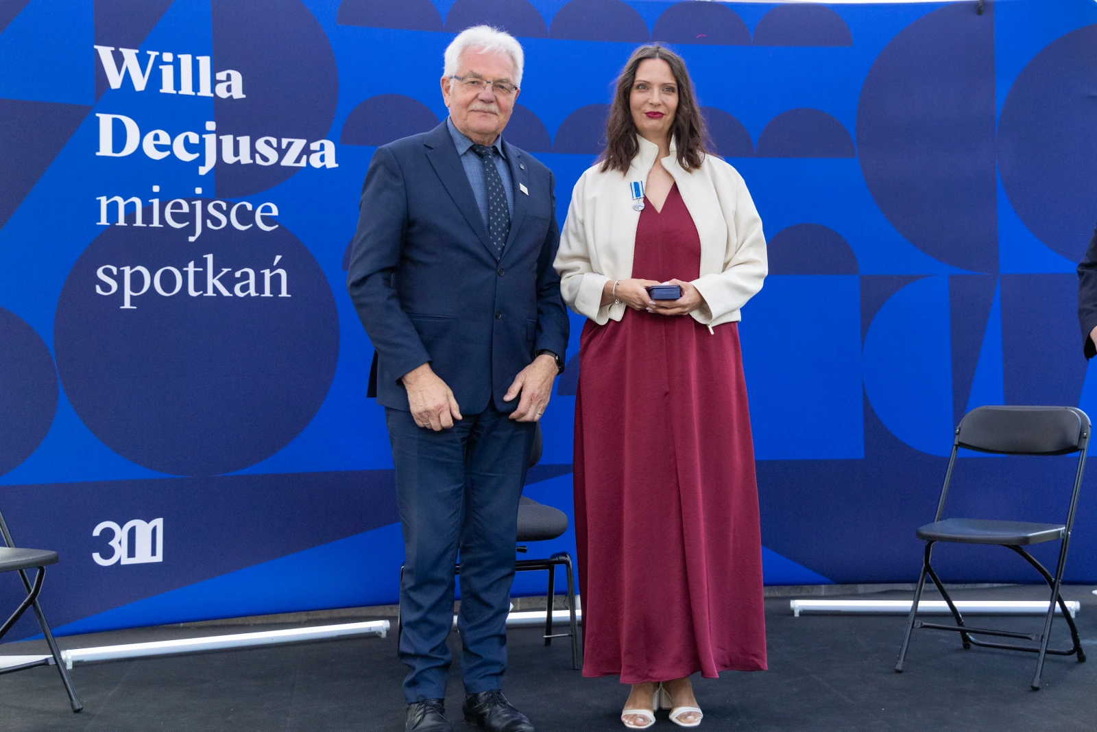 Uroczystość wręczenia odznaczeń w Willa Decjusza. Dwie osoby na tle niebieskiej ścianki.  Autor: Bogusław Świerzowski