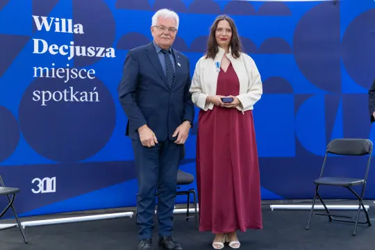 Uroczystość wręczenia odznaczeń w Willa Decjusza. Dwie osoby na tle niebieskiej ścianki.