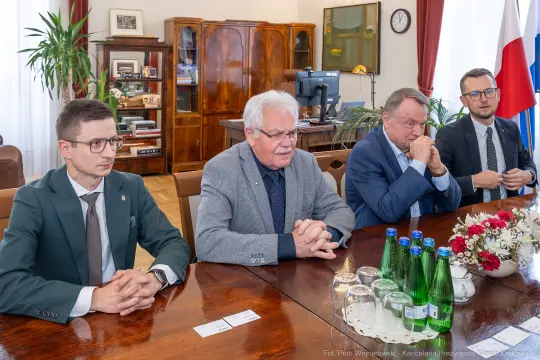 Czterech mężczyzn w garniturach siedzi przy stole, na którym znajdują się butelki wody i kwiaty.
