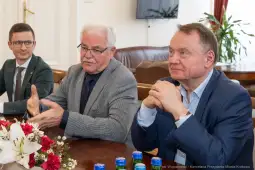 Trzech mężczyzn siedzących przy stole, rozmawiających w formalnej atmosferze.