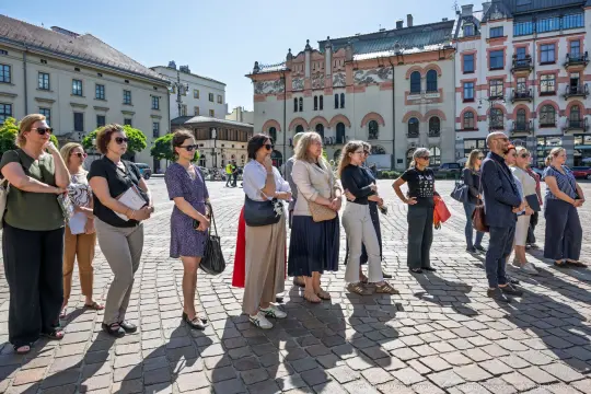 Grupa ludzi stojąca na placu, w tle architektura i słoneczne niebo.