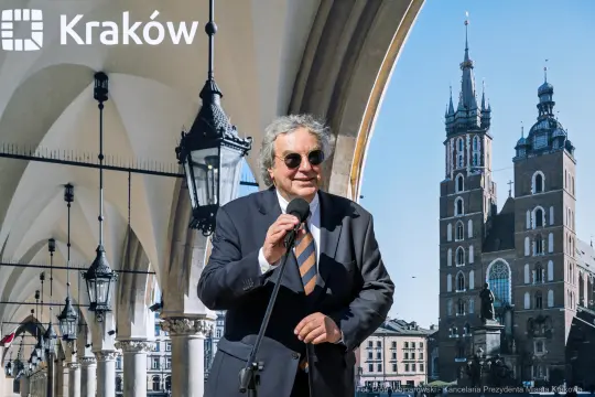 Mężczyzna w okularach przeciwsłonecznych trzyma mikrofon, w tle wieże Kościoła Mariackiego.