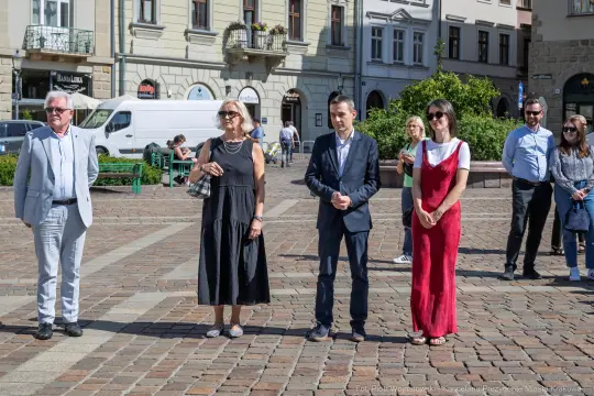 Cztery osoby stojące na placu, w tle budynki i zieleń.