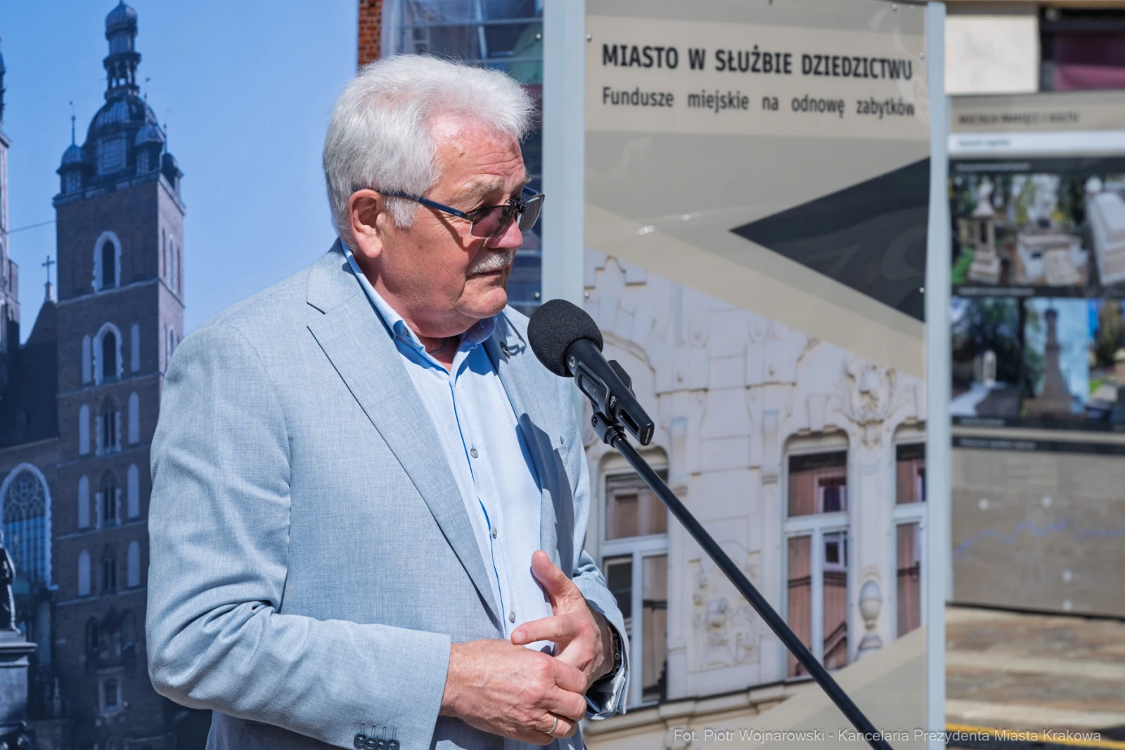 Mężczyzna w jasnym garniturze przemawia do mikrofonu na tle plakatów informacyjnych.  Autor: Piotr Wojnarowski